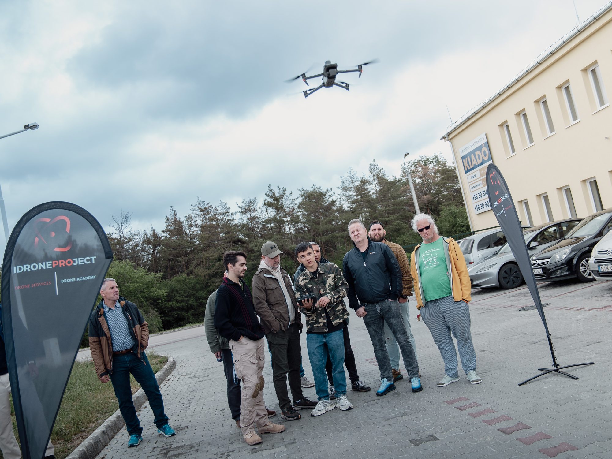 Csoport nézi a levegőben lévő drónt egy korábbi iDroneProject képzésen, az iDroneProject bannerrel